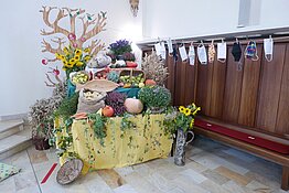 Der Tisch ist geschmückt und gefüllt mit Obst, Gemüse, Getreide und Blumen und, auf einer Leine Mundschutzmasken. 