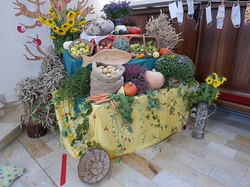 Der Tisch ist geschmückt und gefüllt mit Obst, Gemüse, Getreide und Blumen und, auf einer Leine Mundschutzmasken. 