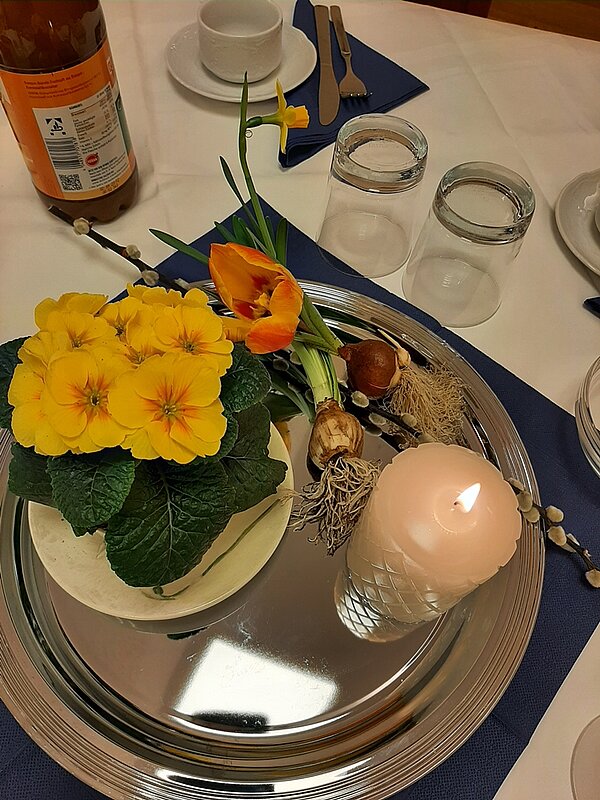 Auf einem silbernen Tablett ein Blumentopf mit gelben Primeln, Tupenzwiebeln mit einer blühenden Tupe, Weidenkätzchen und einer weißen brennenden Kerze